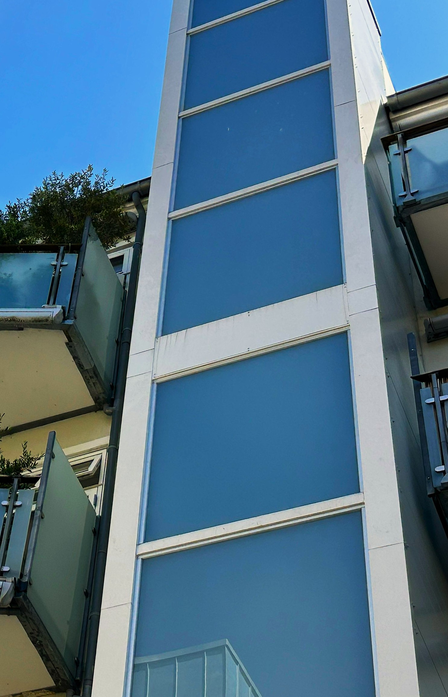 Tall white-framed section of a blue glass-building facade under a clear blue sky, with glass balconies visible on the left.