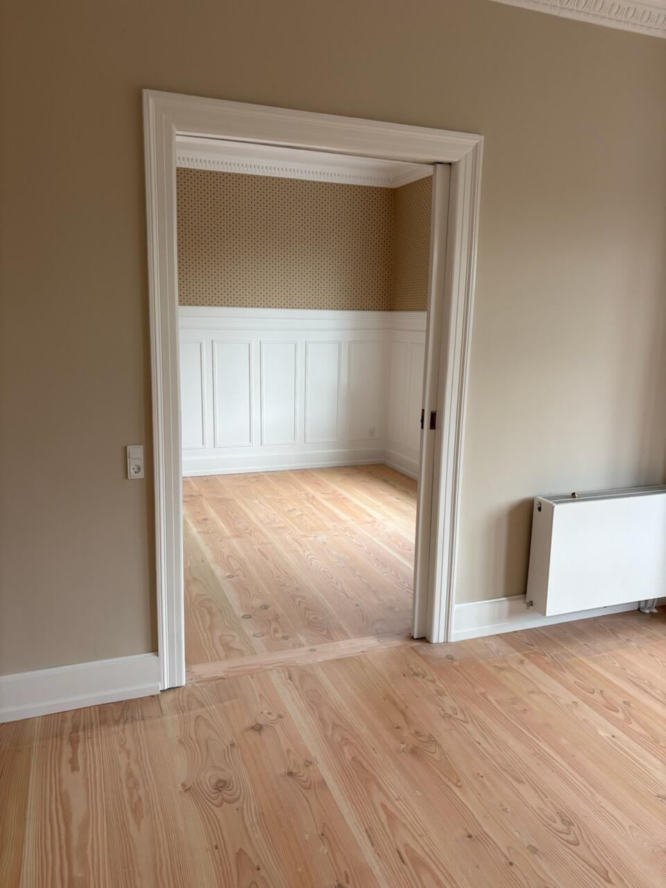 Interior doorway opening into a sunlit room with light wood floors, beige walls, and white wainscoting; radiator visible on the right side of the doorway.